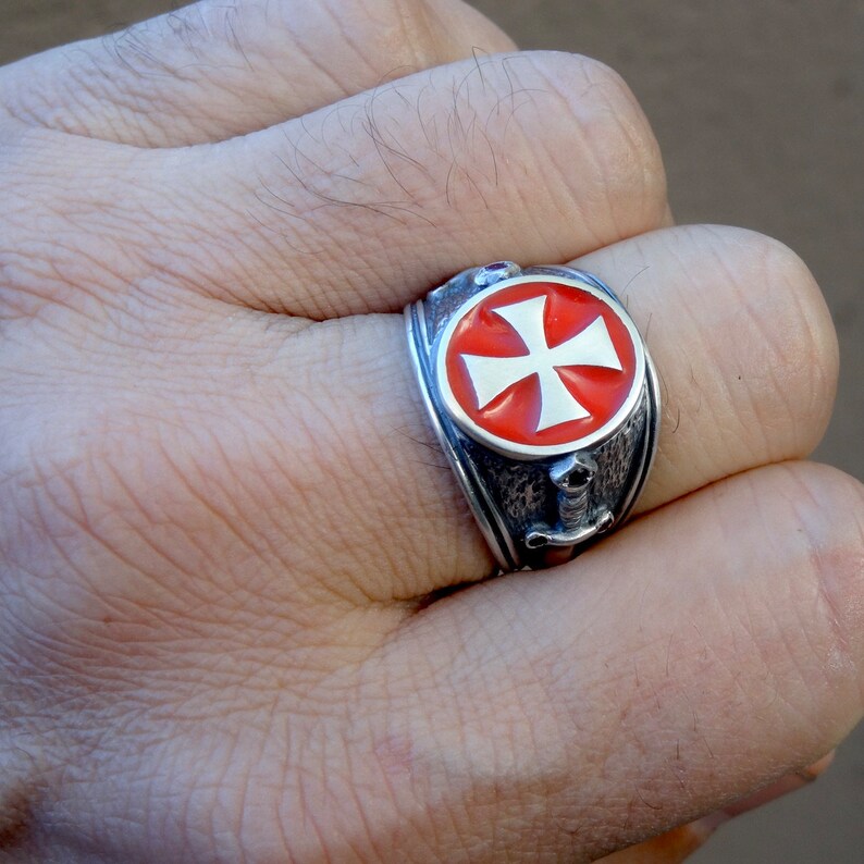Puede incluir: Un anillo de plata con una cruz de esmalte rojo y detalles de espada. El anillo se lleva en un dedo.