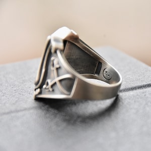 May include: A silver ring with a detailed design featuring a sword and a letter L. The ring is sized 7.