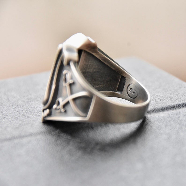 May include: A silver ring with a detailed design featuring a sword and a letter L. The ring is sized 7.