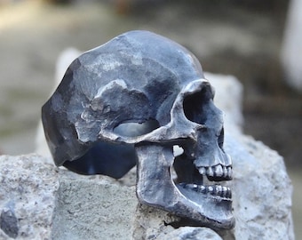 May include: A silver skull ring with a detailed design. The ring is made of metal and has a textured finish.
