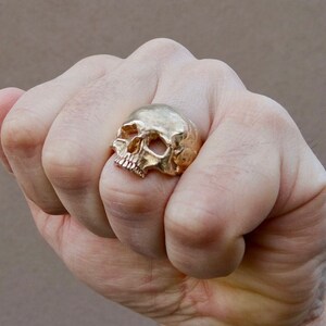 May include: A gold skull ring with a detailed design. The ring is worn on the index finger of a hand.