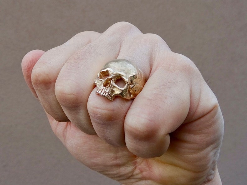 May include: A gold skull ring with a detailed design. The ring is worn on the index finger of a hand.