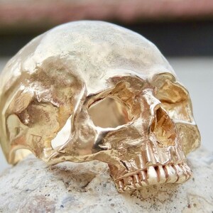 May include: A gold-colored skull ring with detailed features, including a prominent brow ridge, eye sockets, and teeth. The ring is made of metal and has a realistic skull design.