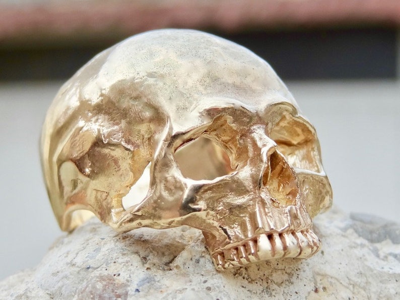May include: A gold-colored skull ring with detailed features, including a prominent brow ridge, eye sockets, and teeth. The ring is made of metal and has a realistic skull design.