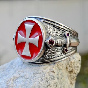 Puede incluir: Un anillo de plata con una cruz de esmalte rojo y un dise&ntilde;o de espada. El anillo tiene tres peque&ntilde;as piedras preciosas rojas.