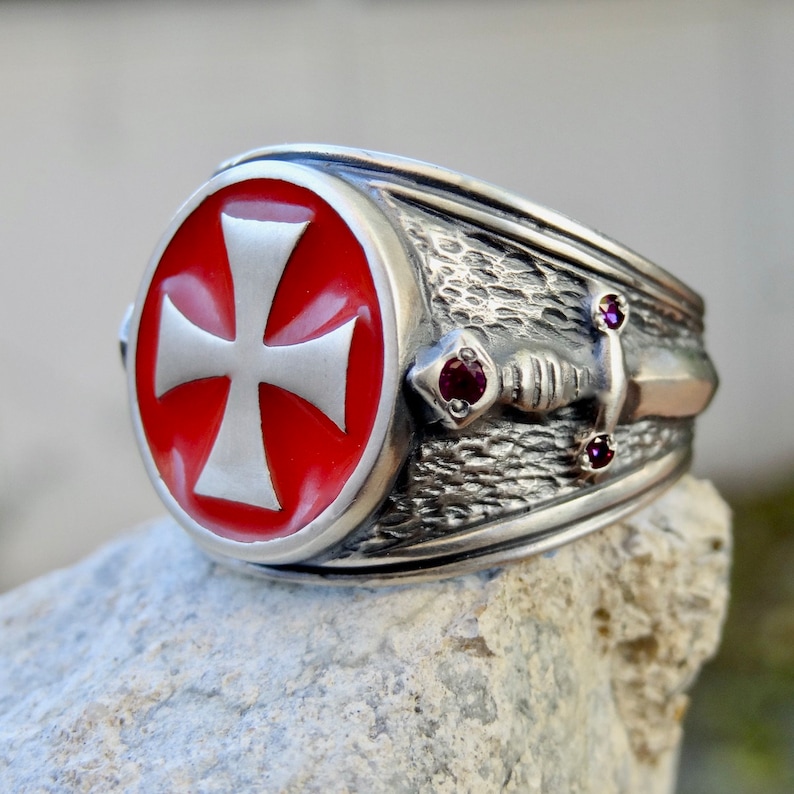 Puede incluir: Un anillo de plata con una cruz de esmalte rojo y un dise&ntilde;o de espada. El anillo tiene tres peque&ntilde;as piedras preciosas rojas.