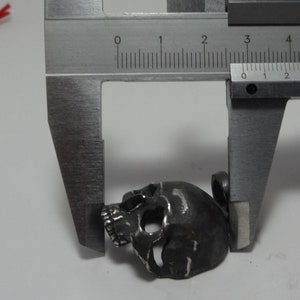 May include: A silver skull pendant charm measured with a caliper. The skull is approximately 2 cm wide.