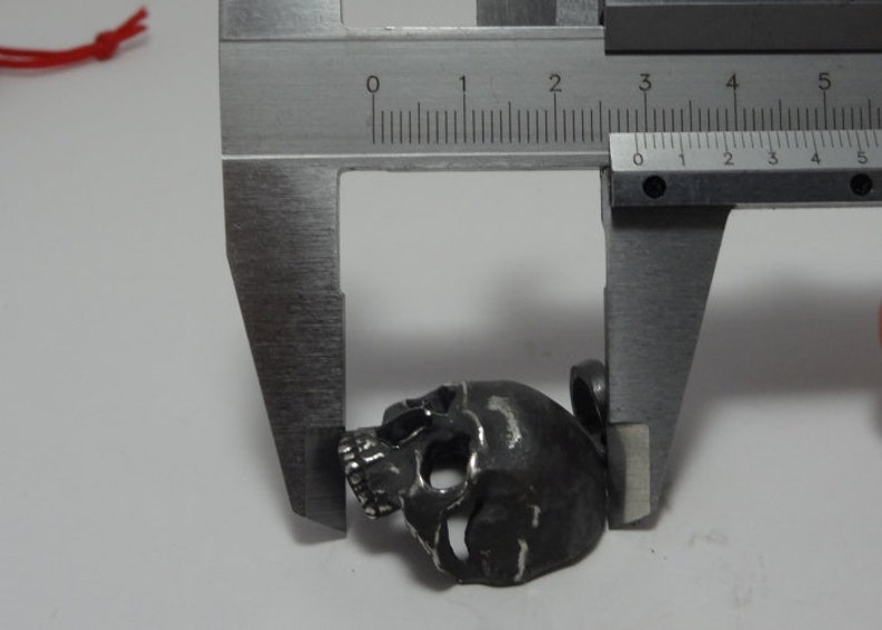 May include: A silver skull pendant charm measured with a caliper. The skull is approximately 2 cm wide.