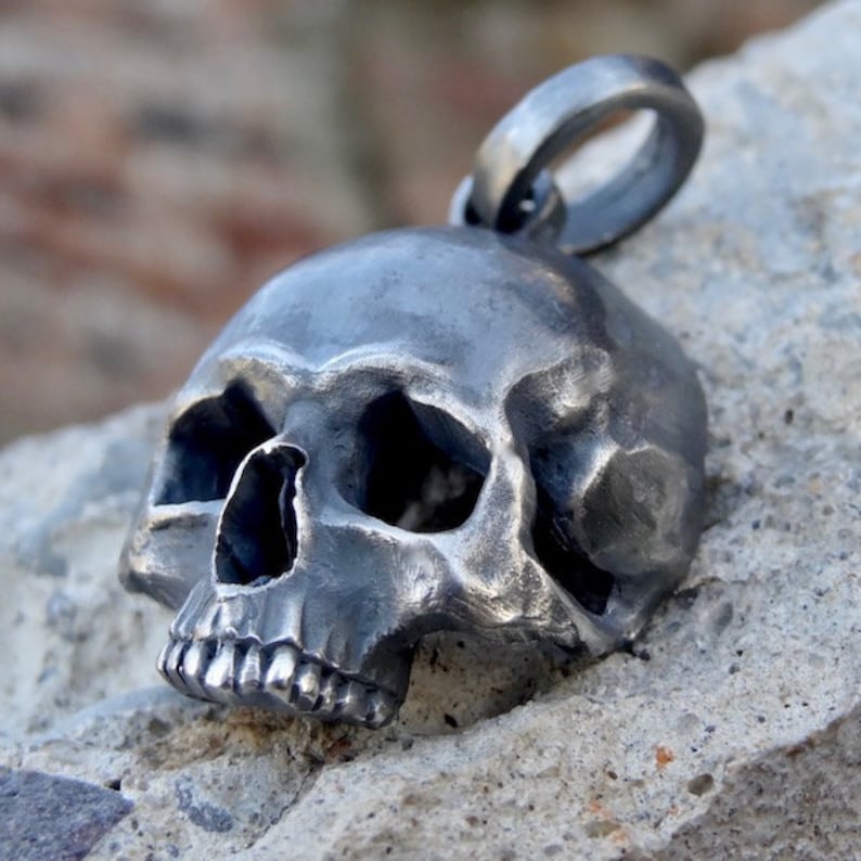 May include: A detailed silver skull pendant with a textured surface. The skull has a prominent jawline and visible teeth. The pendant has a loop at the top for attaching to a chain.