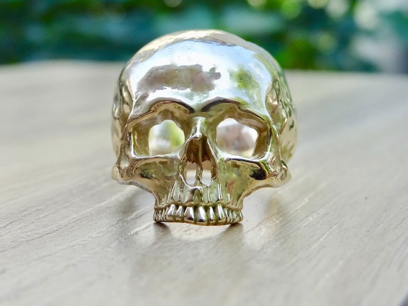 May include: A silver skull ring with a detailed design. The ring is made of metal and has a smooth, polished finish.