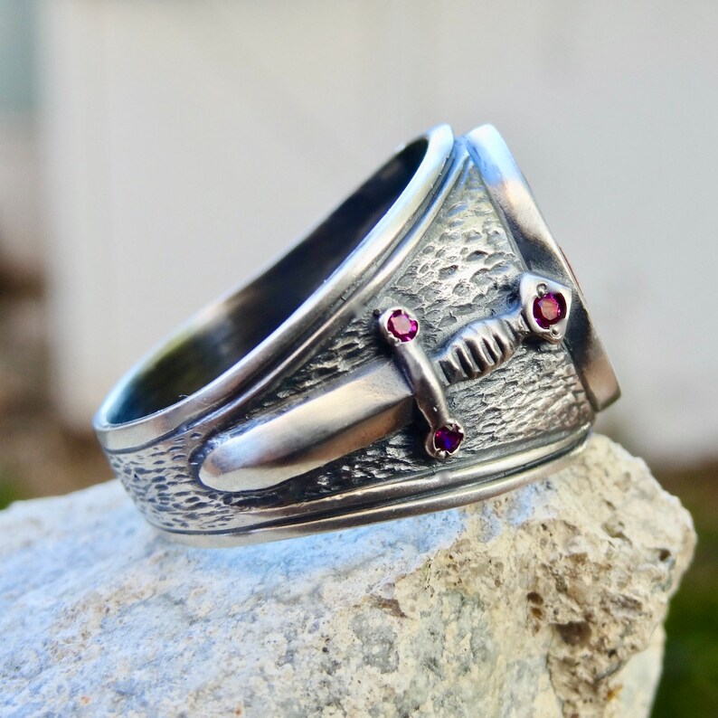 Puede incluir: Un anillo de plata con una banda texturizada y un dise&ntilde;o de espada. La espada tiene tres peque&ntilde;as piedras preciosas rojas.