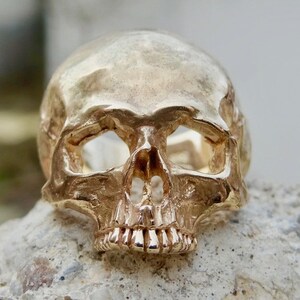 May include: A gold-colored skull ring with a detailed design. The ring is made of metal and has a realistic skull design.