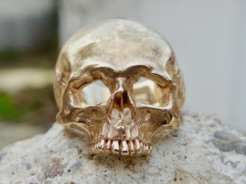 May include: A gold-colored skull ring with a detailed design. The ring is made of metal and has a realistic skull design.