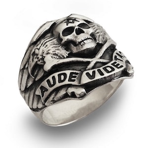May include: A silver ring with a skull and crossbones design. The skull has a pentagram on its forehead and the words "AUDE VIDERI" are inscribed on a banner.