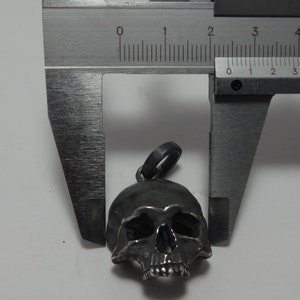 May include: A silver skull pendant charm with a detailed design, measured with a caliper to be approximately 2.5 cm in length.