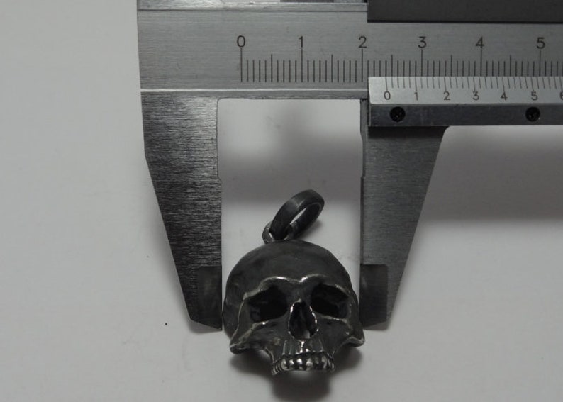May include: A silver skull pendant charm with a detailed design, measured with a caliper to be approximately 2.5 cm in length.