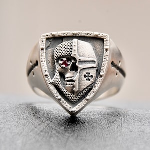 Könnte beinhalten: Ein silberner Ring mit einem schildförmigen Design, das einen Schädel zeigt, der einen Helm mit einem Kreuz trägt. Der Schädel hat einen roten Edelstein als Auge.