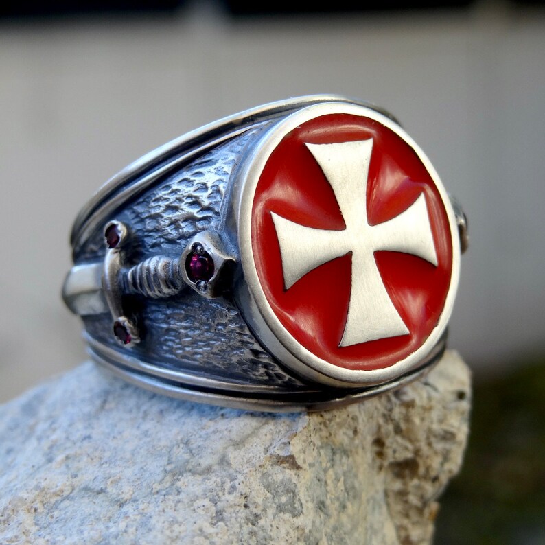 Puede incluir: Un anillo de plata con una cruz de esmalte rojo y un dise&ntilde;o de espada. El anillo tiene tres peque&ntilde;as piedras preciosas rojas.