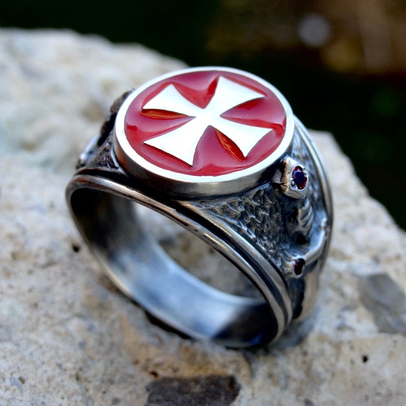Puede incluir: Un anillo de plata con una cruz de esmalte rojo y dos piedras de color rub&iacute; en la banda.
