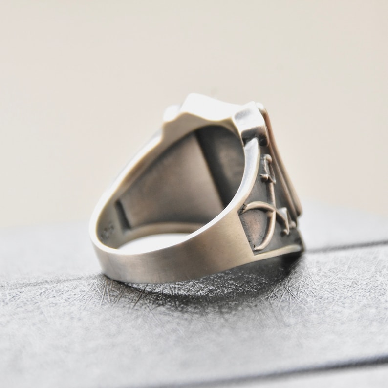 May include: A silver ring with a square face and intricate designs on the band. The ring is on a gray surface.