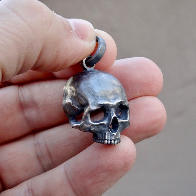 May include: A silver skull pendant with a detailed design. The skull is slightly tilted to the side, showing the eye sockets and teeth.