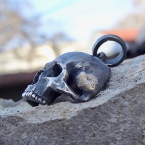 May include: A silver skull pendant with a detailed design. The skull is facing to the side and has a ring attached to the top for hanging.