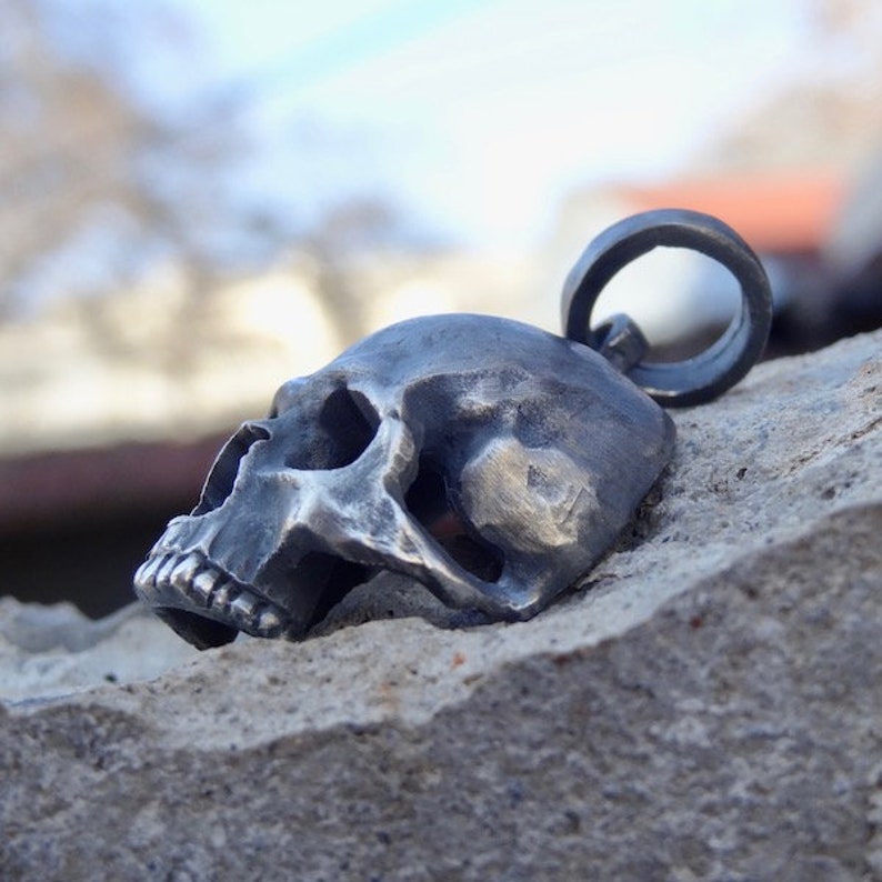 May include: A silver skull pendant with a detailed design. The skull is facing to the side and has a ring attached to the top for hanging.