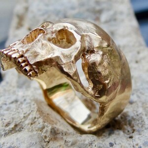 May include: A gold skull ring with a detailed design. The ring is made of solid gold and has a textured finish.