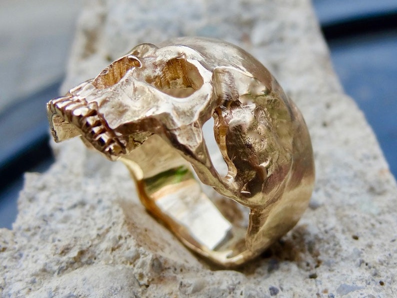 May include: A gold skull ring with a detailed design. The ring is made of solid gold and has a textured finish.