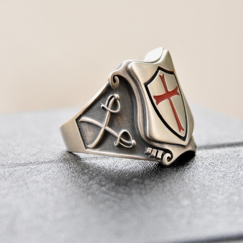 May include: A silver ring with a shield design featuring a red cross. The shield is surrounded by a decorative border with two swords crossed behind it.