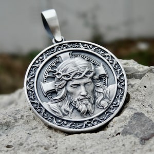 Pode incluir: Pingente de prata com um relevo detalhado de Jesus Cristo com uma coroa de espinhos, rodeado por um design ornamentado.