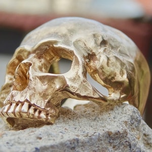 May include: A gold skull ring with a detailed design. The skull is depicted with a prominent jawline and teeth, and the eye sockets are hollowed out.