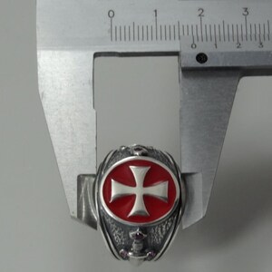 Puede incluir: Un anillo de plata con una cruz de esmalte rojo y un dise&ntilde;o detallado. El anillo se est&aacute; midiendo con un calibre.