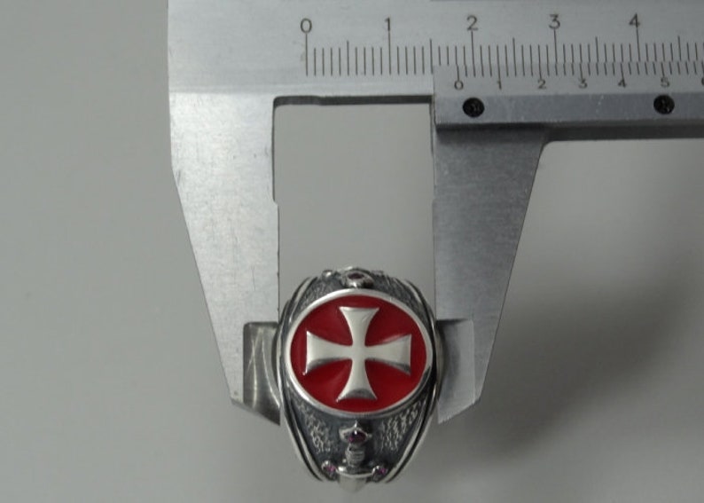 Puede incluir: Un anillo de plata con una cruz de esmalte rojo y un dise&ntilde;o detallado. El anillo se est&aacute; midiendo con un calibre.