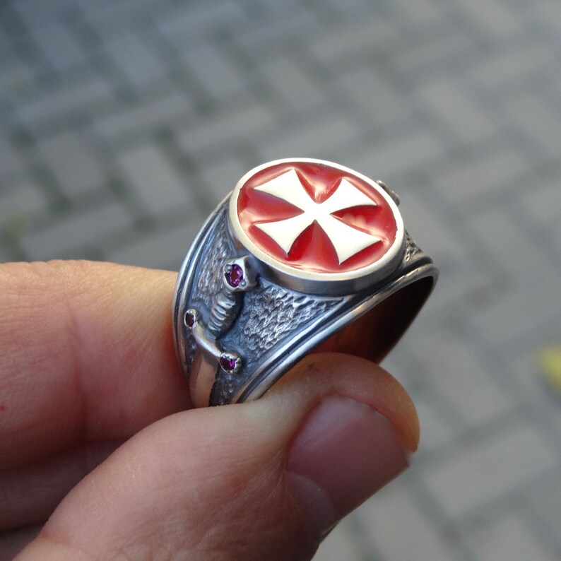 Puede incluir: Un anillo de plata con una cruz de esmalte rojo y dos peque&ntilde;as piedras preciosas rojas. El anillo tiene una banda texturizada y un dise&ntilde;o detallado.