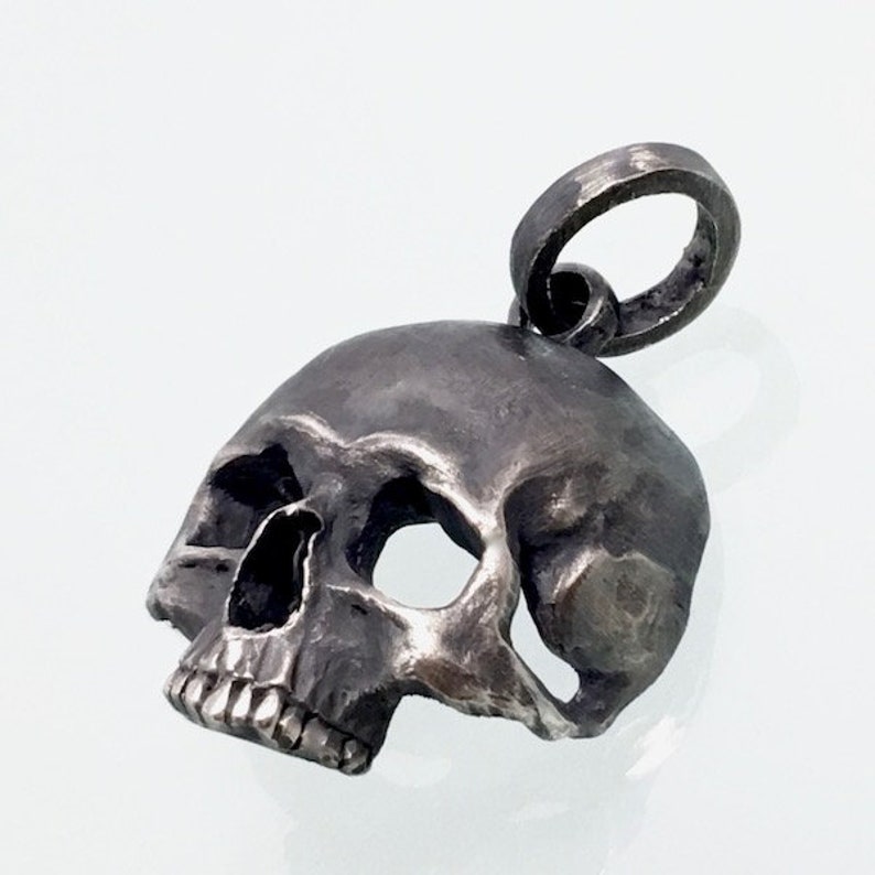May include: A silver skull pendant with a detailed design. The skull has a textured surface and a loop at the top for attaching to a chain.