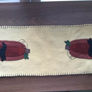 May include: A cream-colored felt table runner with a stitched border. Two appliqued pumpkins with black birds are centered on the runner.