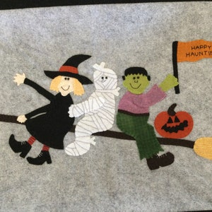May include: A felt Halloween wall hanging with a witch, mummy, and Frankenstein monster riding a broomstick. The witch is wearing a black dress and a pointed hat. The mummy is wrapped in white bandages. The Frankenstein monster is wearing green pants and a purple shirt. The broomstick has a yellow handle and a brown stick. There is a pumpkin and a flag that says "Happy Haunting" in the background.