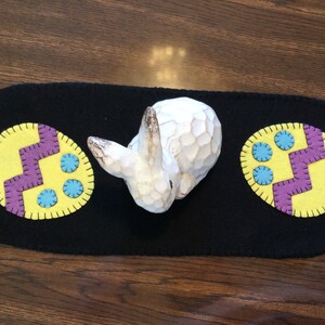 May include: A black felt table runner with two yellow felt Easter eggs with purple and blue accents. A white wooden bunny is in the centre of the runner.