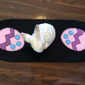 Puede incluir: Un camino de mesa de fieltro negro con dos huevos de Pascua de fieltro rosa y morado con detalles de fieltro azul. Un conejito de madera blanco se sienta en el centro del camino de mesa.