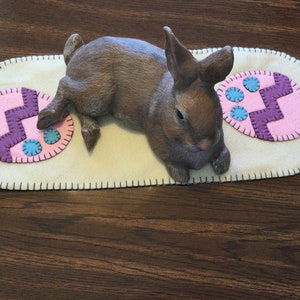 Puede incluir: Una figurita de conejo marrón tumbada sobre un camino de mesa de fieltro blanco con diseños de huevos de Pascua rosas y morados. El camino de mesa tiene un borde cosido negro.