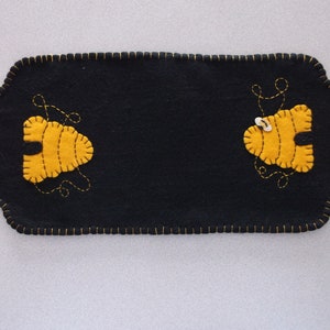 Puede incluir: Caminero de mesa de fieltro negro con dos aplicaciones de fieltro amarillo en forma de abeja. Las abejas tienen ojos negros y un botón blanco para el cuerpo.