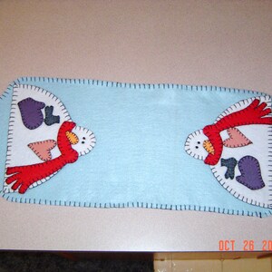 Puede incluir: Un camino de mesa de fieltro azul con dos muñecos de nieve bordados. Cada muñeco de nieve tiene una bufanda roja, ojos negros y una nariz de zanahoria. Los muñecos de nieve se miran.