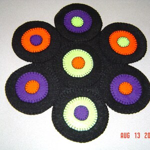 Può includere: Un sottobicchiere a forma di fiore in feltro nero con sette cerchi. Ogni cerchio ha un cerchio più piccolo all'interno con colori alternati di arancione, viola e verde.