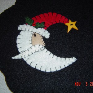 May include: A felt appliqué of a white crescent moon with a red and white Santa hat, a green holly sprig, and a yellow star on a black background.