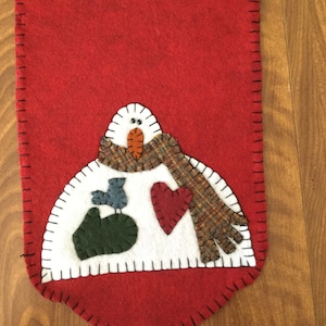 Puede incluir: Un camino de mesa de fieltro rojo con un diseño de muñeco de nieve. El muñeco de nieve tiene un cuerpo blanco, una nariz de zanahoria y una bufanda a cuadros. También se incluye un pájaro azul y un corazón rojo. Los bordes están cosidos con hilo negro.