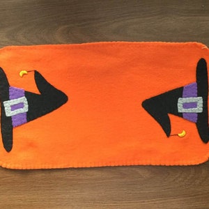 Puede incluir: Un camino de mesa de fieltro naranja con dos sombreros de bruja negros con detalles en morado y gris. Los sombreros tienen medias lunas amarillas en la parte superior.