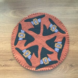 Può includere: Un tappeto rotondo in feltro arancione con un design bianco e nero di cinque cappelli da strega. I cappelli hanno accenti viola e dorati.