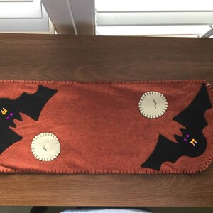 Op de afbeelding: Een felt tafelkleed met een Halloween-thema. Het heeft twee zwarte vleermuizen met paarse ogen en oranje tanden, die over witte felt manen vliegen. Het tafelkleed is roestoranje van kleur met een gestikte rand.