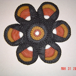 Puede incluir: Una alfombra de fieltro negro en forma de flor con seis pétalos. Cada pétalo tiene un diseño de maíz dulce en naranja, marrón y blanco. El centro de la alfombra tiene un círculo con círculos concéntricos en naranja, marrón y blanco.